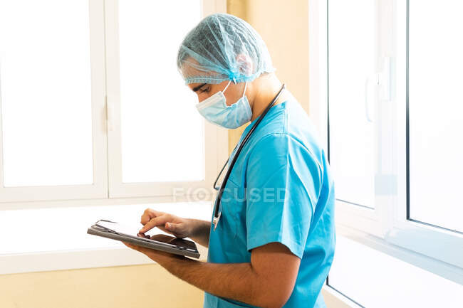 Médico masculino de uniforme e com estetoscópio em pé no hospital e navegar na Internet em tablet — Fotografia de Stock