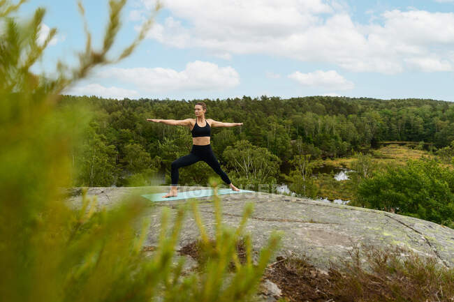 Mujer joven descalza de cuerpo entero haciendo pose de Guerrero sobre alfombra y mirando hacia otro lado mientras practica yoga sobre roca en la naturaleza en verano - foto de stock