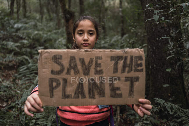 Ethnic kid raising cardboard piece with Save The Planet inscription while looking at camera in green forest — Stock Photo
