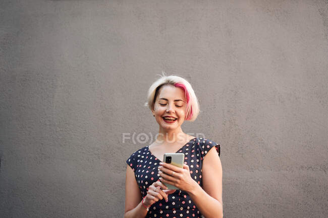 Розсудлива неформальна жінка з коротким волоссям і в одязі повідомлення на мобільному телефоні стоячи проти сірої стіни на вулиці і дивлячись на екран — стокове фото