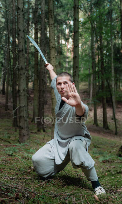 Hombre de cuerpo entero con ropa tradicional practicando la postura de la espada durante el entrenamiento de kung fu en el bosque - foto de stock