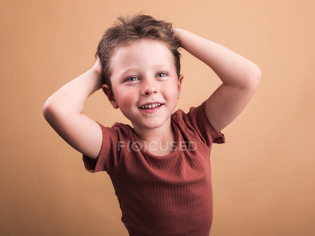 Criança satisfeita em roupas casuais com cabelo castanho olhando para longe com o sorriso dos dentes e com as mãos na cabeça — Fotografia de Stock