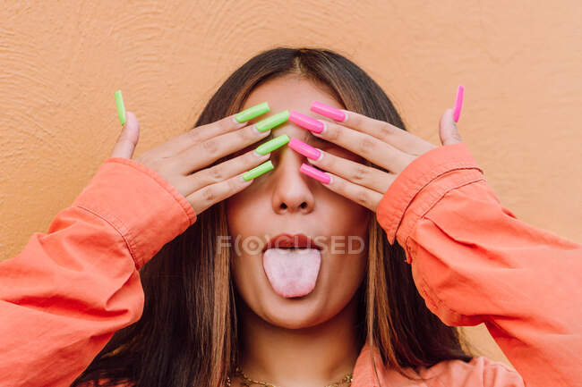 Naughty female showing tongue while covering eyes with hands with long bright nails against orange background — Stock Photo