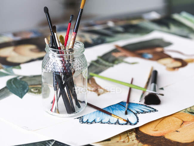 Стеклянная банка с разнообразными кисточками, размещенная на различных творческих картинах на столе в мастерской — стоковое фото