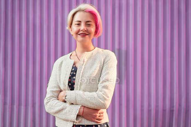 Charming alternative female with dyed hair and in trendy clothes standing against violet wall in city and looking at camera — Stock Photo
