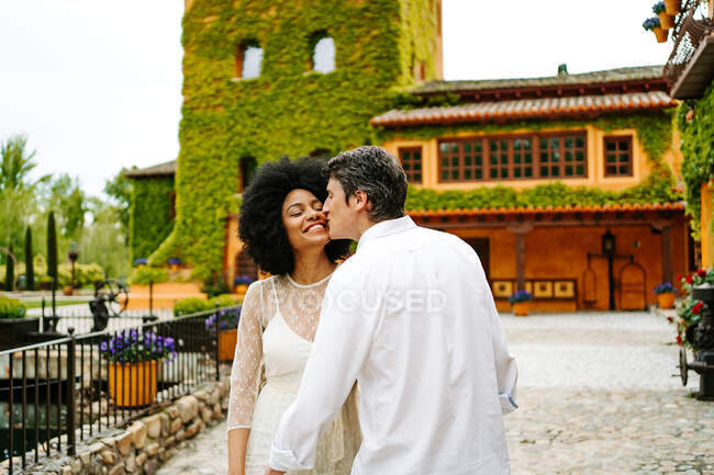 Loving man kissing black woman on cheek while standing near building with ivy on walls in park in summer — Stock Photo