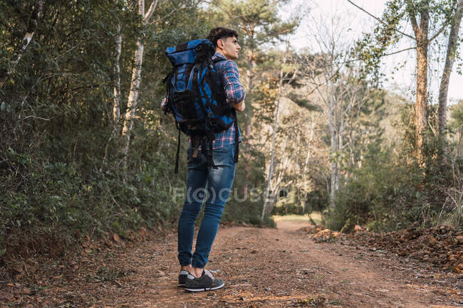 Side view of traveling man with backpack standing on sandy road in forest during trekking and looking away — Stock Photo