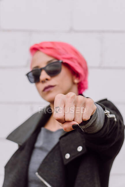 Молодая модная женщина в стильном наряде и солнечных очках показывает кулак с маленькой татуировкой на пальце, стоя рядом с белой стеной на улице — стоковое фото