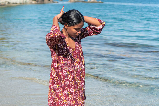 Ethnic female traveler in sundress with floral ornament looking down with hands behind head against ocean and rock — Stock Photo