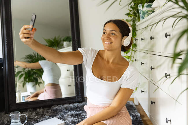 Mulher étnica encantadora em fone de ouvido sem fio tomando auto-retrato no celular enquanto sentado no tapete entre espelho e laptop em casa — Fotografia de Stock