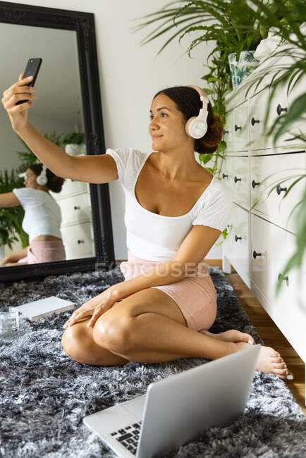 Charmante ethnische Frau in drahtlosem Headset macht Selbstporträt auf dem Handy, während sie zu Hause auf dem Teppich zwischen Spiegel und Laptop sitzt — Stockfoto