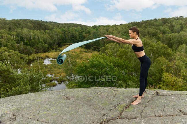 Ganzkörper-Barfußläuferin in schwarzer Aktivkleidung rollt Matte auf Felsen zu Beginn der Yoga-Session in der Nähe von Sumpf in der Natur aus — Stockfoto