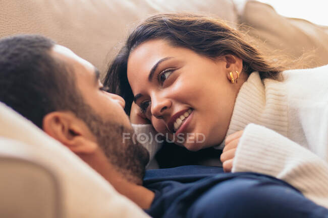 Amante mulher hispânica étnica em suéter deitado no homem no sofá em abraço enquanto olham um para o outro — Fotografia de Stock