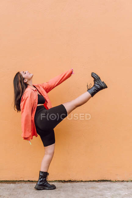 Vista lateral cuerpo completo de alegre milenial femenino en traje elegante pateando el aire contra la pared naranja - foto de stock