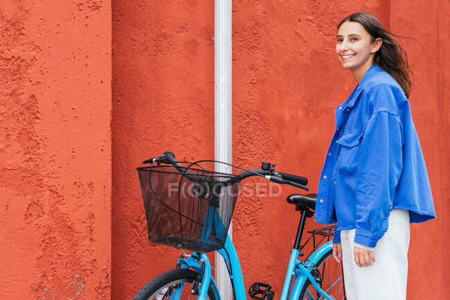 Vista lateral da fêmea encantada em pé perto da bicicleta moderna com cesta na rua e olhando para longe — Fotografia de Stock