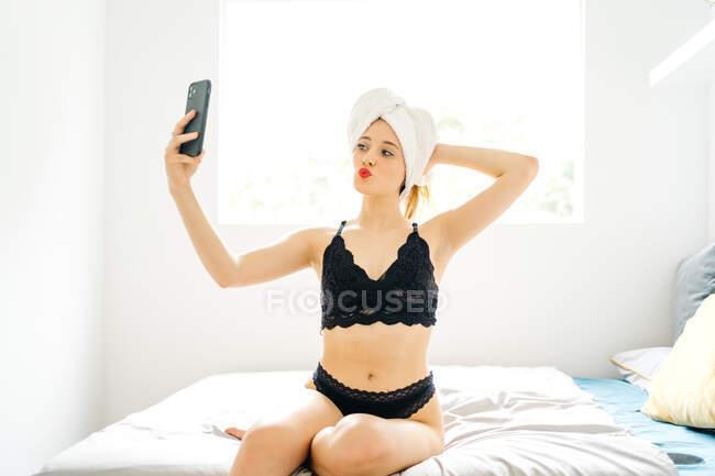 Charmante Frau im Handtuch-Turban und in schwarzer Dessous, die rote Lippen schmollt und Selbstaufnahmen auf dem Smartphone macht, während sie zu Hause im Bett sitzt — Stockfoto