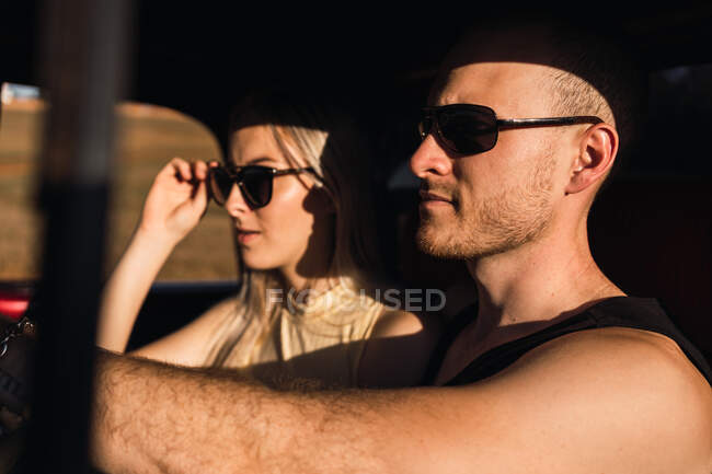 Fidanzato fresco seduto in auto con ragazza cool in occhiali da sole nella giornata di sole — Foto stock