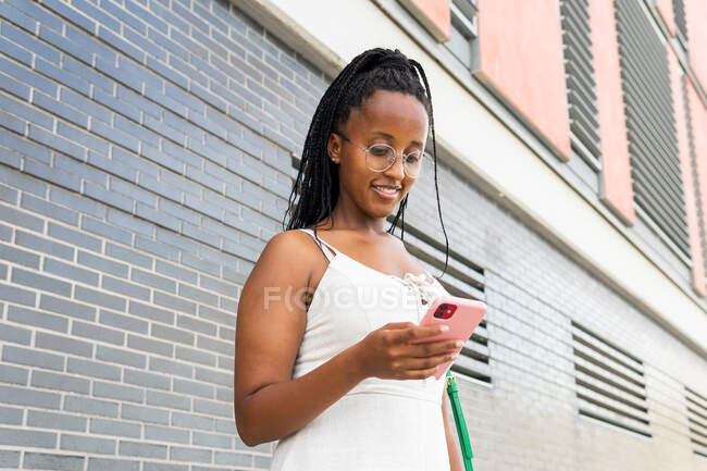 Angle bas de sourire afro-américaine avec des messages sur les médias sociaux via téléphone portable tout en se tenant dans la rue avec de vieux bâtiments à Barcelone — Photo de stock