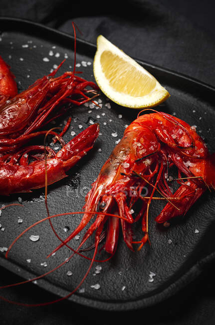 Délicieuses crevettes rouges cuites sur plateau avec du sel grossier et des morceaux de citron juteux sur fond sombre — Photo de stock