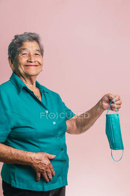 Donna anziana deliziata in piedi con maschera medica blu usa e getta da COVID e guardando la fotocamera su sfondo rosa in studio — Foto stock