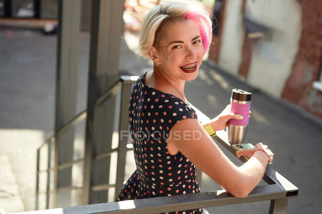 High angle side view of joyful alternative female with dyed hair and in braces standing on stairs with reusable cup of hot takeaway drink and looking at camera — Stock Photo