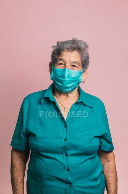 Femme âgée souriante utilisant un masque médical de protection bleu du coronavirus tout en regardant la caméra sur fond rose en studio — Photo de stock