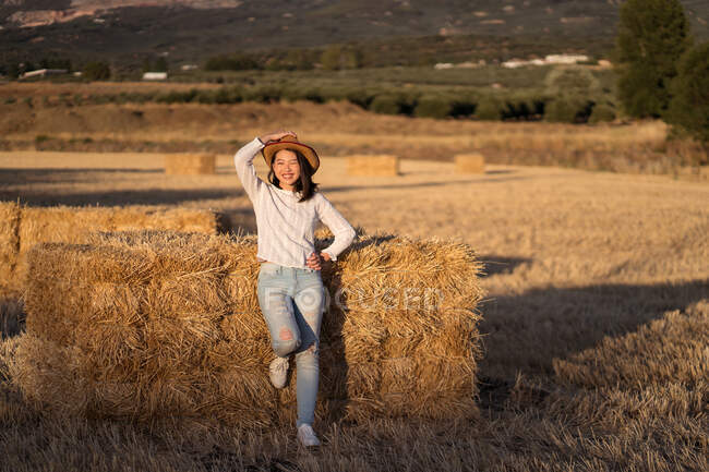 Conteúdo fêmea étnica com chapéu de palha em pé perto da pilha de feno seco no campo e olhando para a câmera — Fotografia de Stock