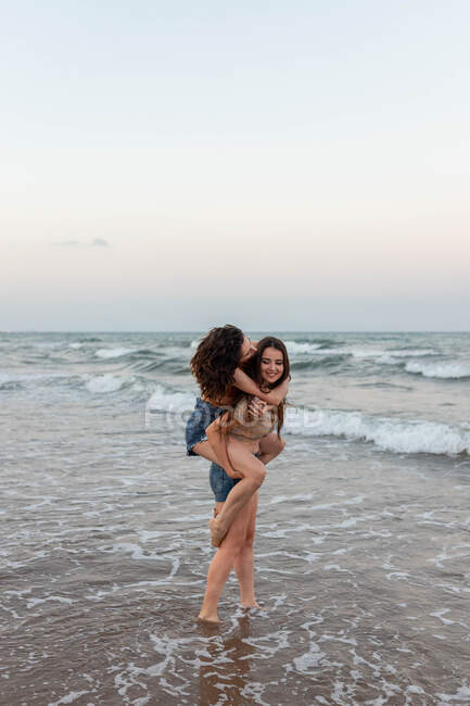 Young woman giving piggyback ride to girlfriend while standing in waving sea water in evening — Stock Photo