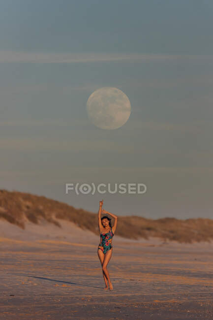 Slim female in swimsuit standing with raised arms on sandy beach in evening under blue sunset sky with moon — Stock Photo