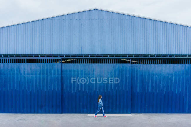 Vista remota da fêmea em vestuário jeans andando ao longo da rua no fundo do edifício azul — Fotografia de Stock