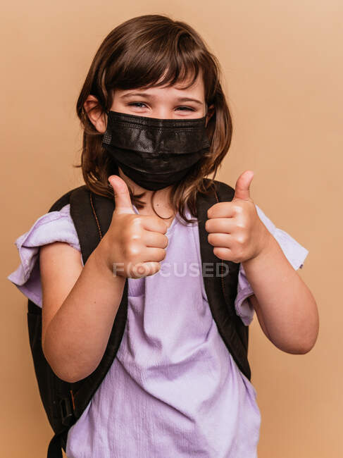 Зачарована дитина з рюкзаком і в захисній масці від коронавірусу, що показує як знак на коричневому фоні в студії і дивиться на камеру — стокове фото
