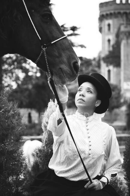 Femme noire adulte sérieuse en chapeau et tenue élégante avec des gants caressant cheval brun près des plantes vertes et des arbres dans la campagne — Photo de stock