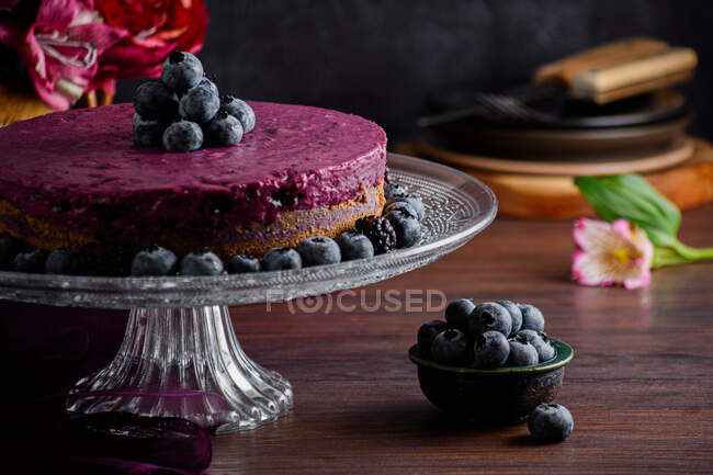 Вкусный черничный торт из мусса с фиолетовым кремом, украшенный свежими ягодами, подаваемый на стеклянном столике на темном столе с цветами — стоковое фото