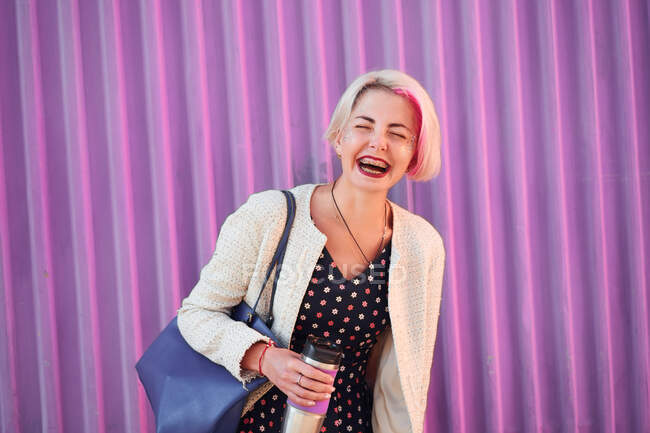 Positive informelle Frau mit gefärbten kurzen Haaren steht mit Getränk in umweltfreundlicher Tasse auf dem Hintergrund der lila Wand in der Stadt und geschlossenen Augen — Stockfoto