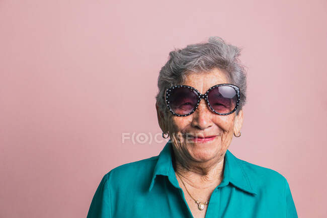 Felice donna moderna invecchiata con i capelli grigi e in occhiali da sole alla moda su sfondo rosa in studio e guardando la fotocamera — Foto stock