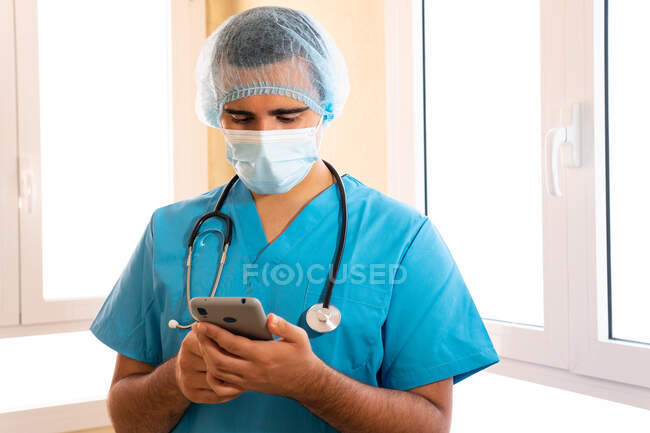 Чоловік у формі і стетоскоп стоїть у лікарні і серфінг Інтернету на смартфоні — стокове фото