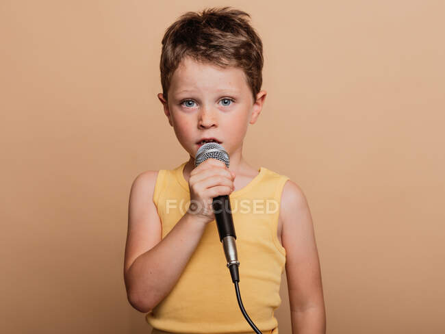 Niño fresco en el canto en micrófono moderno sobre fondo marrón en el estudio - foto de stock