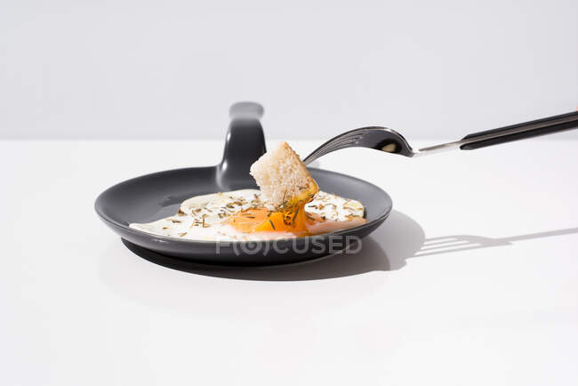 Tenedor de metal con pan fresco sumergido en yema líquida de huevo frito servido en sartén sobre fondo blanco - foto de stock