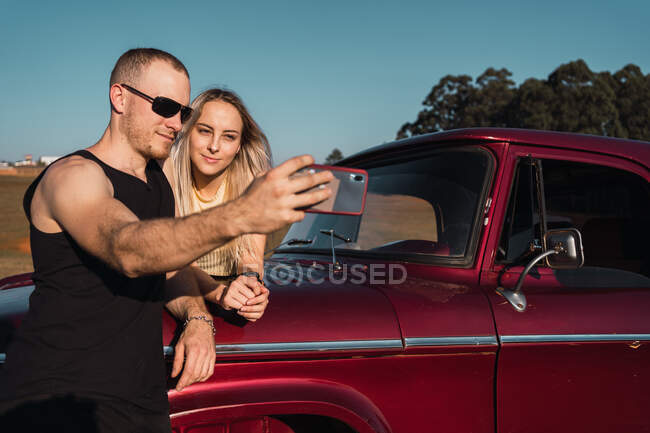 Sorridente coppia cool in piedi vicino rosso auto retrò e prendendo auto colpo su smartphone — Foto stock