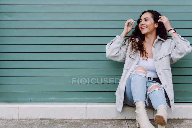 Обаятельная женщина в стильной одежде, сидящая у стены здания, улыбаясь и отводя взгляд — стоковое фото