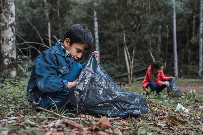 Volontaires ethniques avec des sacs en plastique ramassant les ordures du terrain contre les arbres dans les bois d'été en plein jour — Photo de stock
