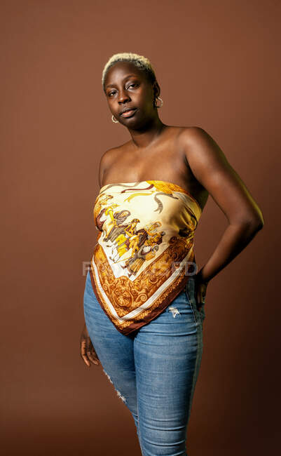 Modèle féminin afro-américain confiant portant un haut en écharpe de soie sur fond brun en studio et regardant la caméra — Photo de stock
