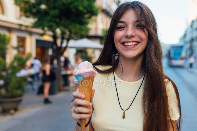 Cortar alegre jovem fêmea em pingente e brincos com delicioso gelato em cone waffle olhando para a câmera na rua — Fotografia de Stock