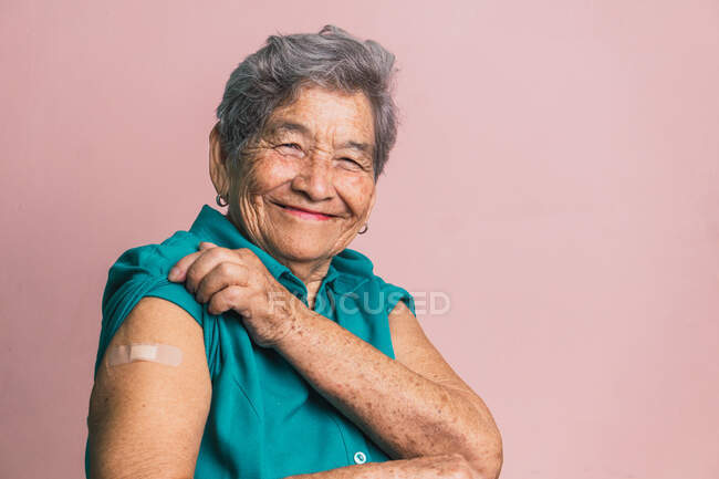 Mulher sênior positiva mostrando braço com patch após a vacinação de COVID em fundo rosa e olhando para a câmera — Fotografia de Stock