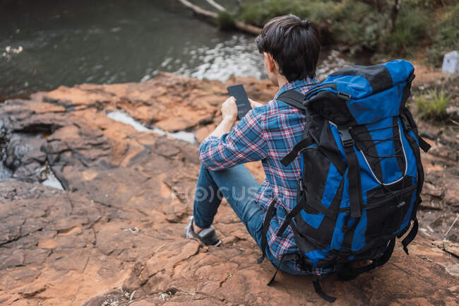 Назад вид содержания мужчина турист с рюкзаком сидя возле озера в лесу и просматривая мобильный телефон во время путешествия — стоковое фото