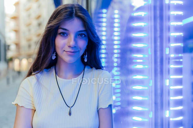Дружелюбная молодая женщина в кулоне и серьгах смотрит в камеру на неоновые лампы в вечернем городе — стоковое фото