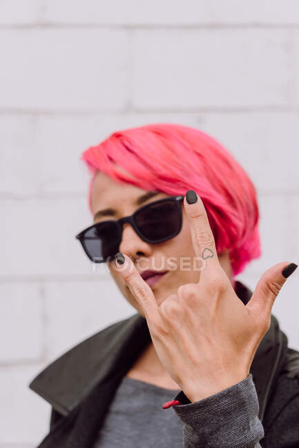 Giovane femmina con capelli rosa brillante in abito alla moda e occhiali da sole che mostrano segno rock and roll su sfondo bianco — Foto stock