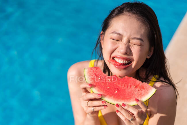 Von oben eine fröhliche ethnische Frau im gelben Bikini, die am Pool sitzt und an einem sonnigen Sommertag mit geschlossenen Augen frische Wassermelone isst — Stockfoto
