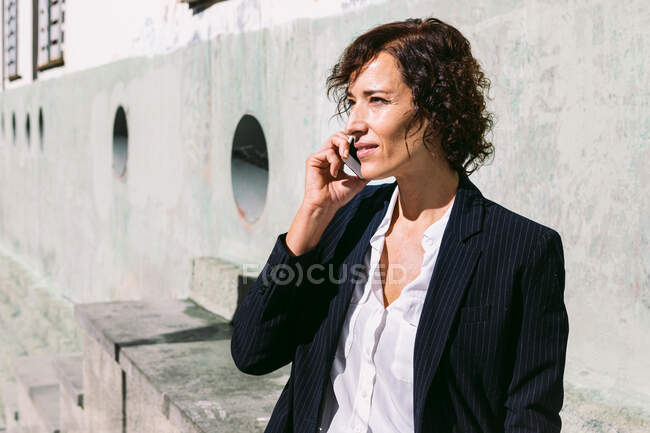 Positivo adulto feminino executivo trabalhador vestindo elegante roupa de pé com a mão na cintura e falando no celular no dia ensolarado — Fotografia de Stock