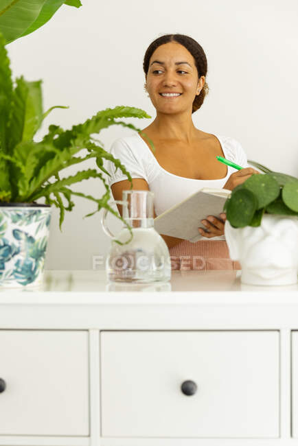 Mulher étnica pensativa com caneta e agenda entre plantas verdes em vasos no jardim da casa — Fotografia de Stock